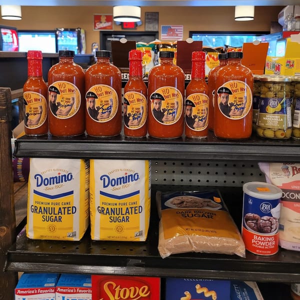 A store shelf displays various hot sauces, Domino sugar, brown sugar, baking powder, and olives.